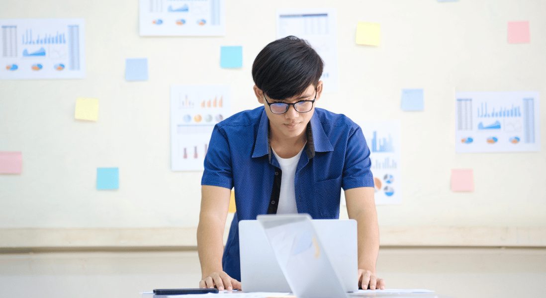 male student , wearing glasses , laptop on the table, board with grades analytics