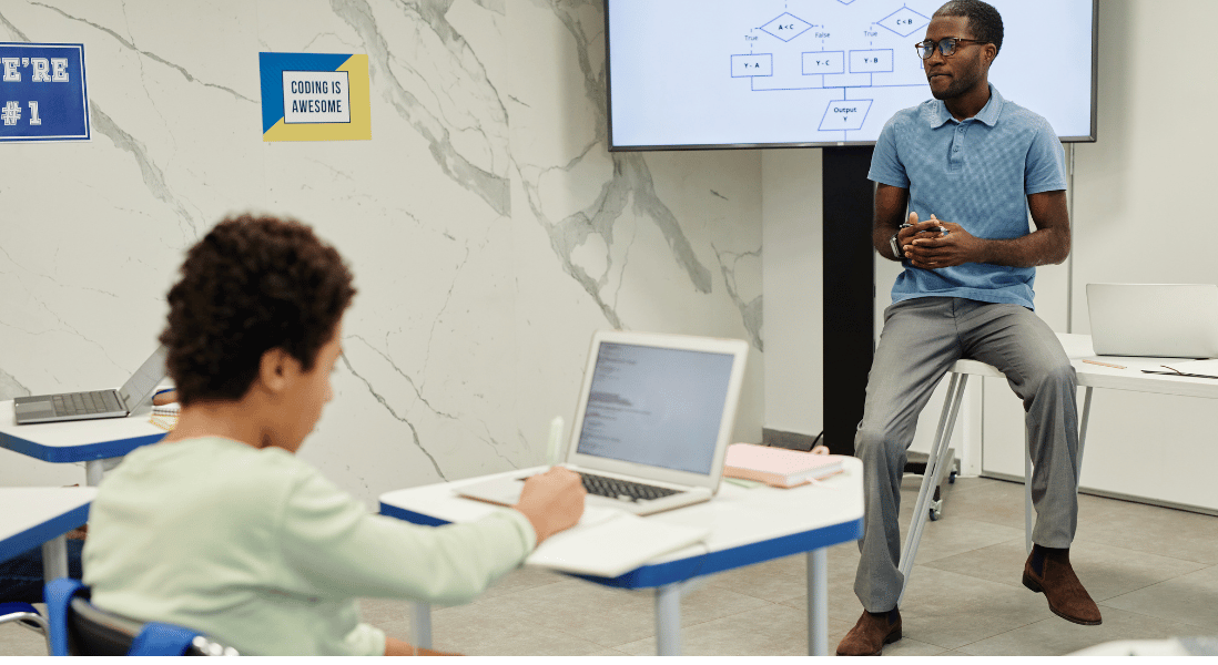 teacher seated on the table in front of the class, male student taking notes on the exercise book, laptop over the table