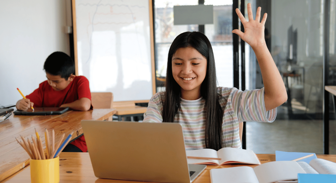 girl raising a left hand , table , laptop , exercise books , pencils , boy in the background