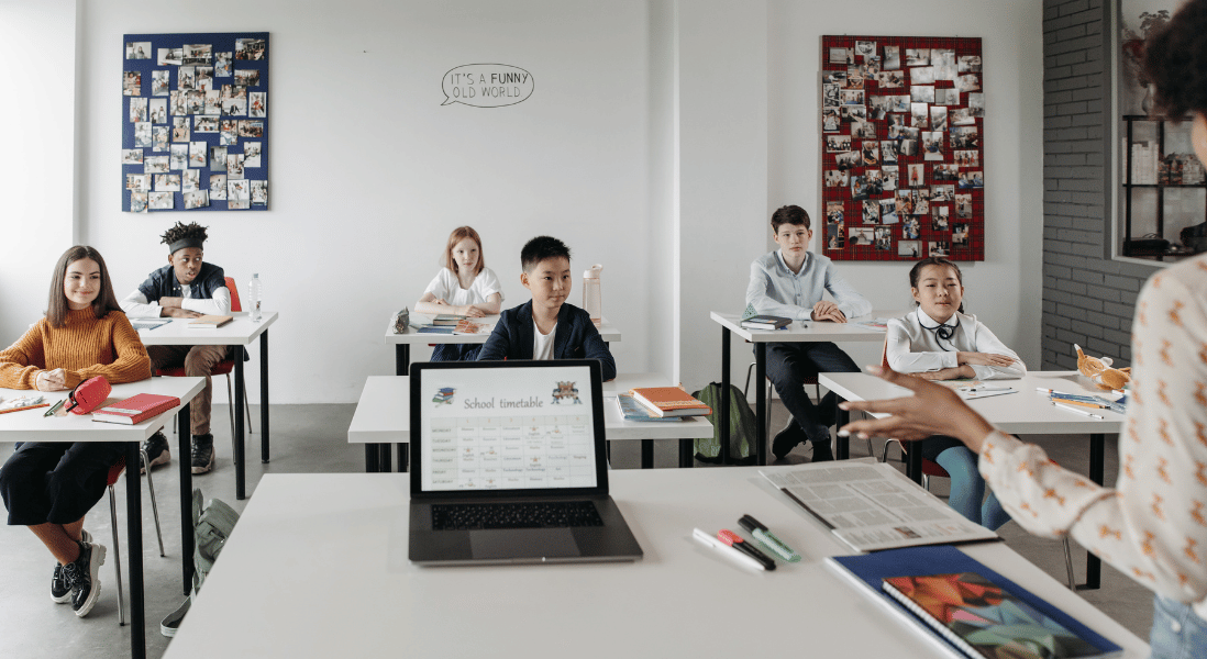 teacher standing in front of the classroom next to her desk , open laptop , learners 