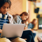 black girl leaning next to the red face brick building , laptop , white headphones