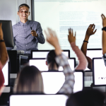 male standing in from of the room , group of people raising their hands ,