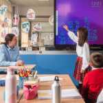 class teacher seated in front of the classroom , learner writing on the display screen