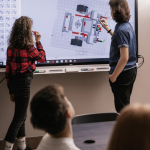2 learners writing on what looks like a white projector sheet other learners seated down